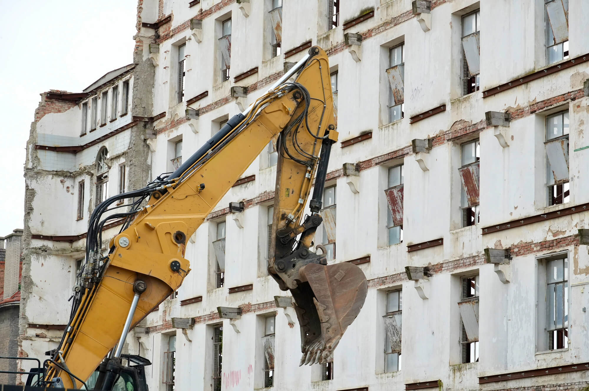 תמונת נושא של המאמר על עורך דין פינוי בינוי המציגה תקריב על כף וזרוע של מחפרון (באגר) צהוב על רקע בניין רב-קומות מתפורר. הבניין בעל קירות לבנים, חלונות ללא זכוכית וסימני הריסה גלויים, כולל קורות תמיכה חשופות וחלקי קיר שבורים.
