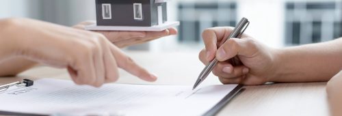 תמונת נושא של המאמר על עורך דין מיסוי מקרקעין המציגה תקריב של ידיים על שולחן עבודה, אחת מצביעה על מקום חתימה במסמך והשנייה חותמת, כשדגם מיניאטורי של בית מוחזק ברקע מעל המסמכים.