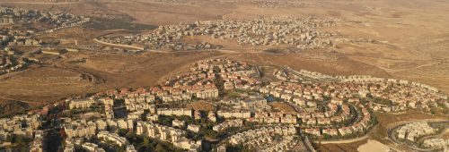 תמונת נושא של המאמר על התחדשות עירונית בירושליים המציגה צילום אווירי של פסגת זאב צפופה המוקפת במדבר או נוף יבש, עם בתים בעלי גגות אדומים ושטחים פתוחים שזורים בכבישים, ויישובים נוספים נראים באופק.