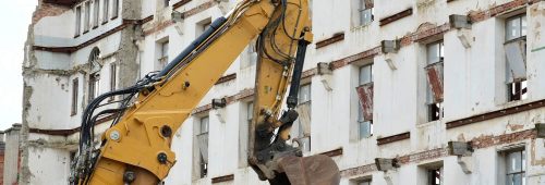 תמונת נושא של המאמר על עורך דין פינוי בינוי המציגה תקריב על כף וזרוע של מחפרון (באגר) צהוב על רקע בניין רב-קומות מתפורר. הבניין בעל קירות לבנים, חלונות ללא זכוכית וסימני הריסה גלויים, כולל קורות תמיכה חשופות וחלקי קיר שבורים.